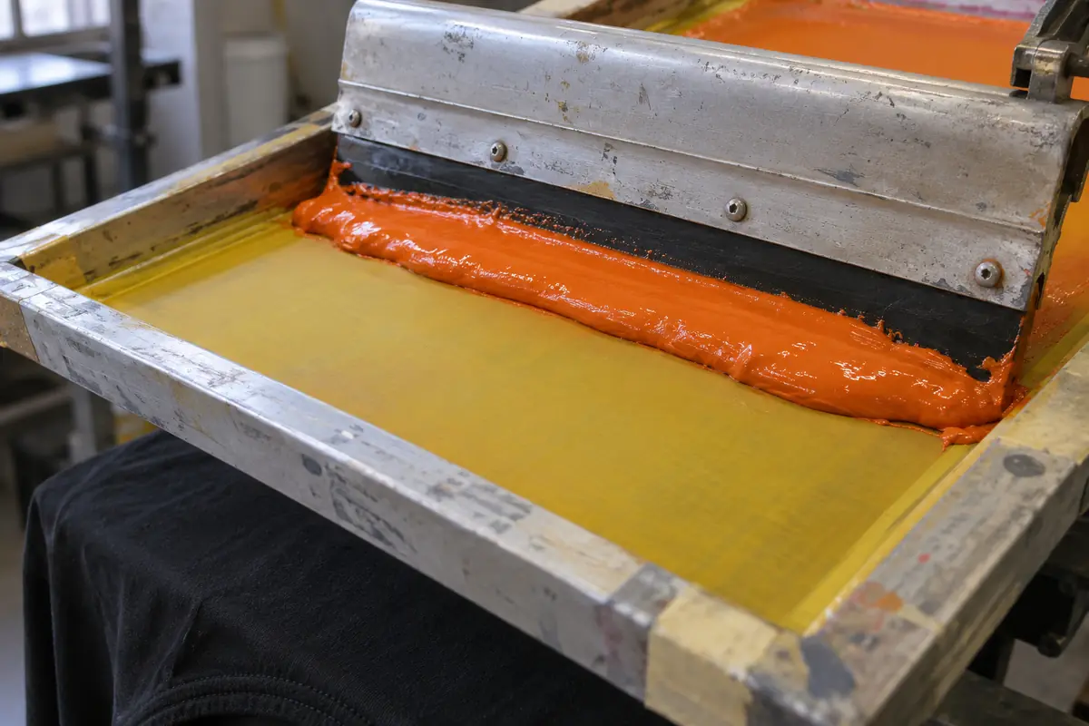 A squeegee pulling orange plastisol ink across a yellow mesh screen